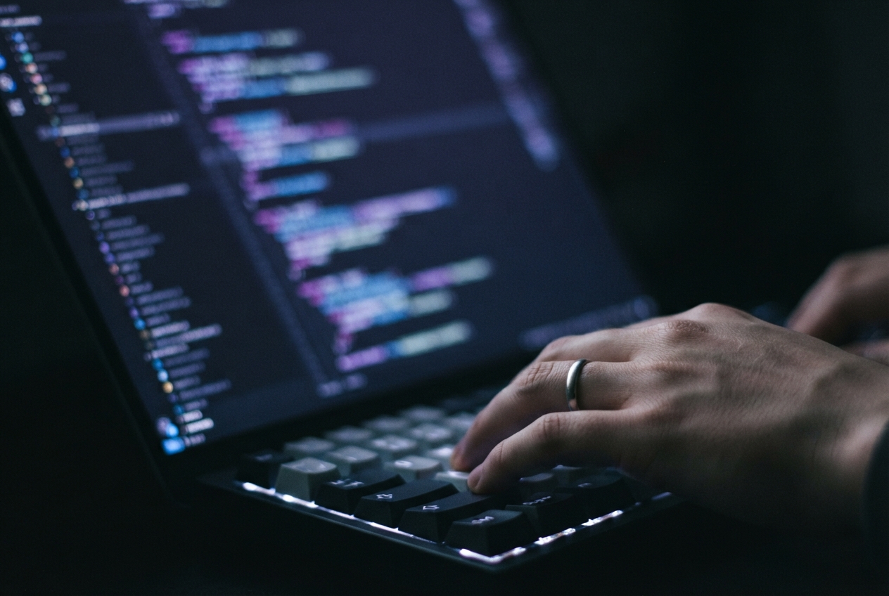 Hand on a backlit keyboard beside a laptop displaying blurred computer code.